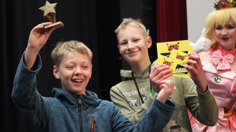 Das Team "Die Dummen" präsentiert stolz den bronzenen Pokal und ihre gewonnenen Gutscheine vom Battle Kart in Bispingen. Im Hintergrund steht eine Cosplayerin als Princess Peach verkleidet.